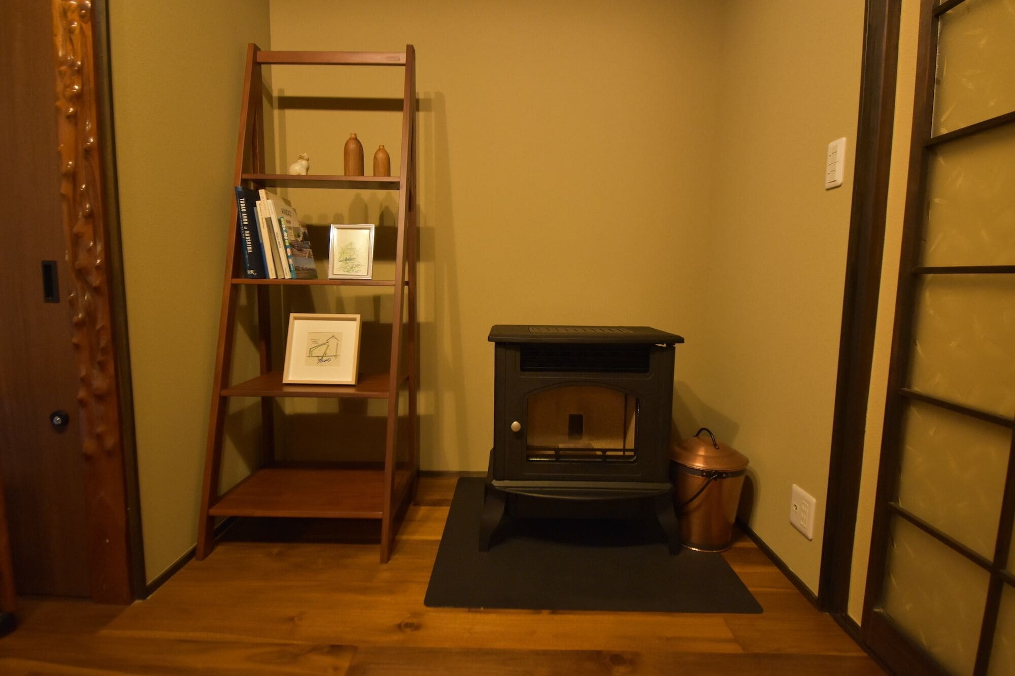 A room with a wooden bookshelf, a black mat on the floor, and a wood-burning stove.