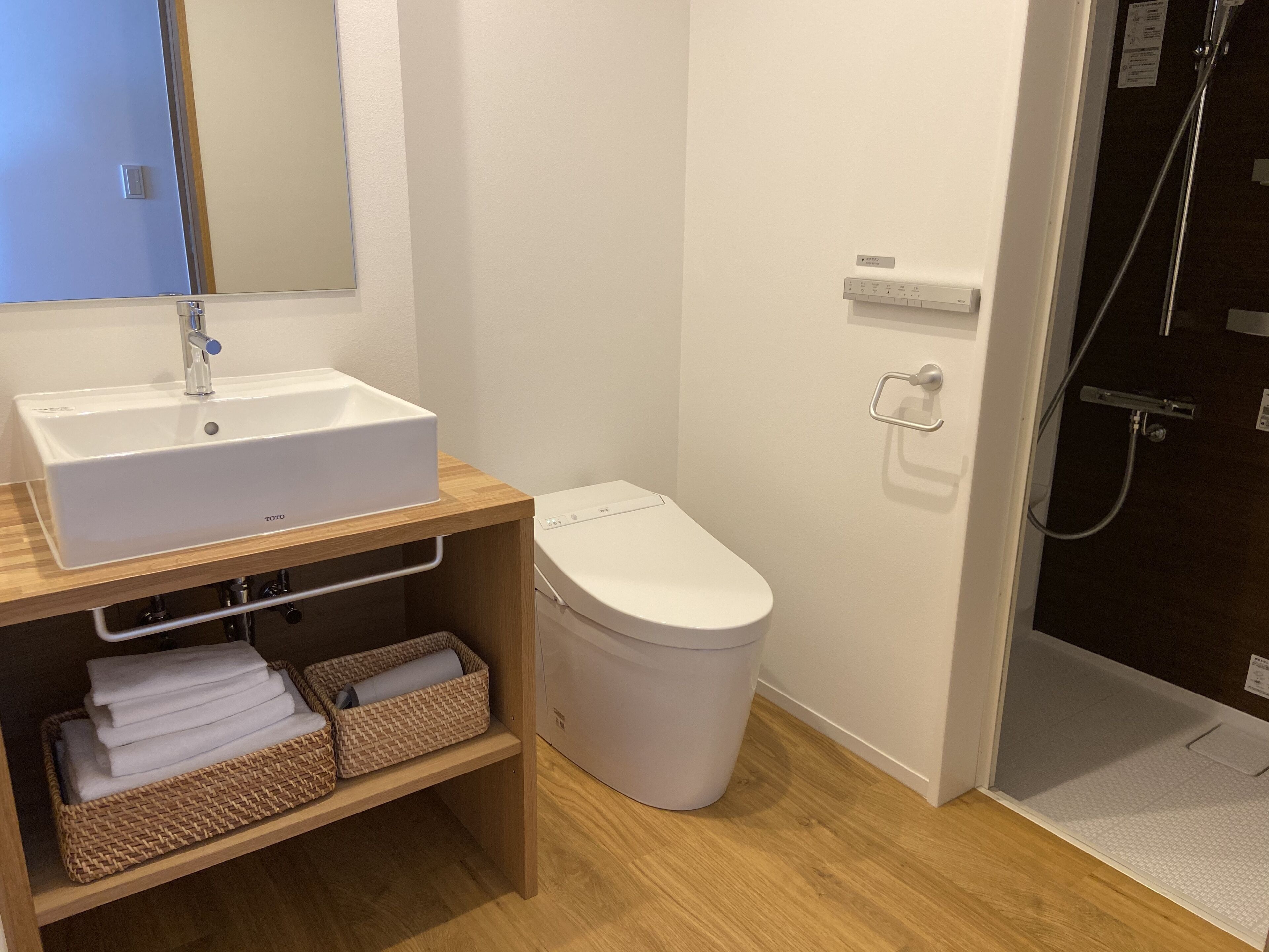 Une salle de bain avec un lavabo, des toilettes et une douche, le tout de couleur blanche. On trouve des paniers en osier sous le lavabo et un miroir au-dessus.