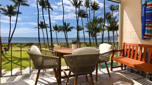 A balcony with wicker chairs and a wooden table, overlooking a beach with palm trees and the ocean. - Oceanfront, Exceptionally Remodeled, Split A/C, King bed, OH the sunsets (Kihei)