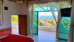 Ein Schlafzimmer mit einem roten Bett, einem Deckenventilator und einem großen Fenster mit Blick auf die Stadt und das Meer.