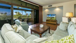 Una sala de estar con sofá, mesa de centro y televisor. Hay un balcón con vistas a palmeras y edificios.