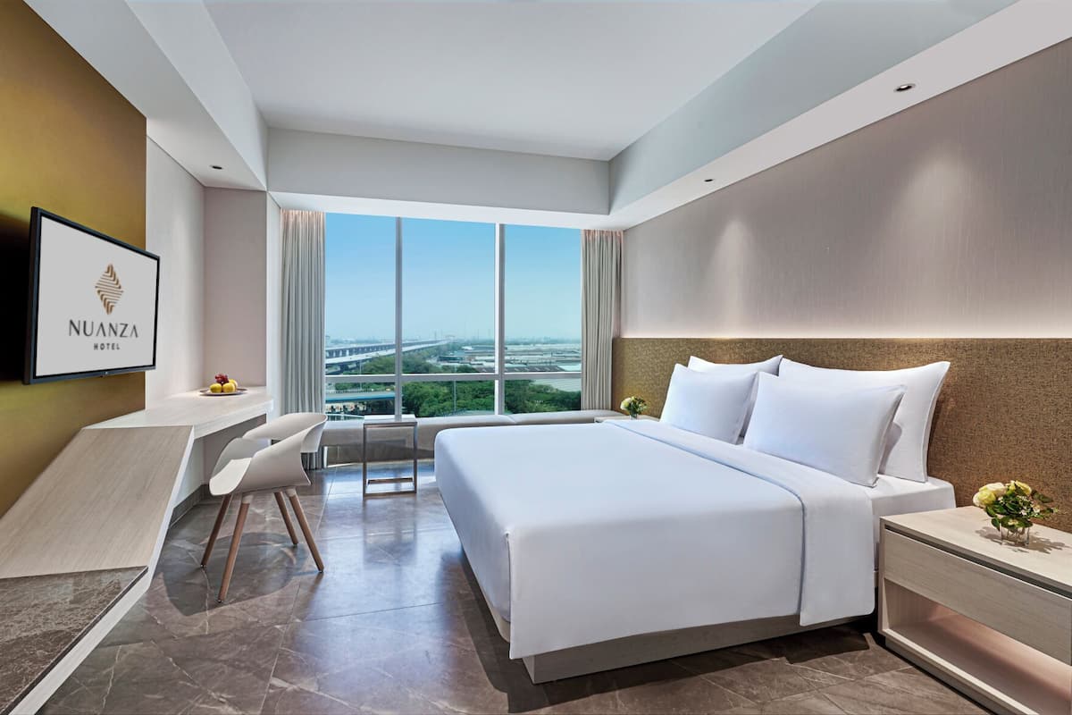Une chambre d’hôtel moderne avec un grand lit, un bureau et une chaise. Il y a un téléviseur au mur et une fenêtre donnant sur la ville.