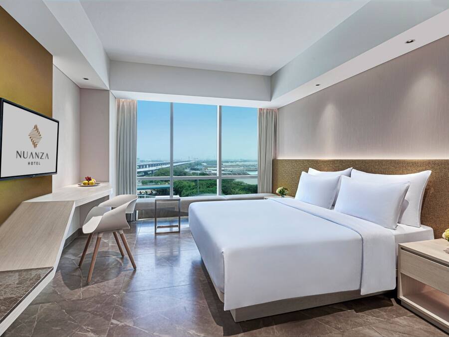 A modern hotel room with a large bed, a desk, and a chair. There is a TV on the wall and a window with a city view.