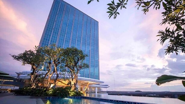 Un edificio moderno con fachada de vidrio reflectante, rodeado de un área ajardinada con árboles y una piscina.