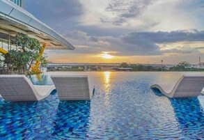 Uma piscina infinita com vista para o pôr do sol, cadeiras de descanso e um edifício moderno ao fundo.