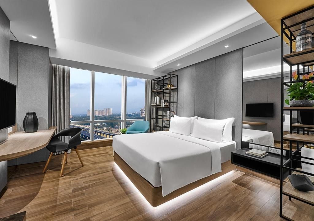 A modern hotel room with a large bed, a desk, a chair, a TV, and a bookshelf.