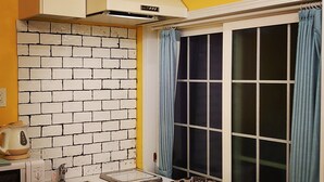 A compact kitchen with a white brick backsplash, a microwave, a kettle, and a patterned chair near a window with blue curtains. - The standard apartment shibuya D (Tokyo)