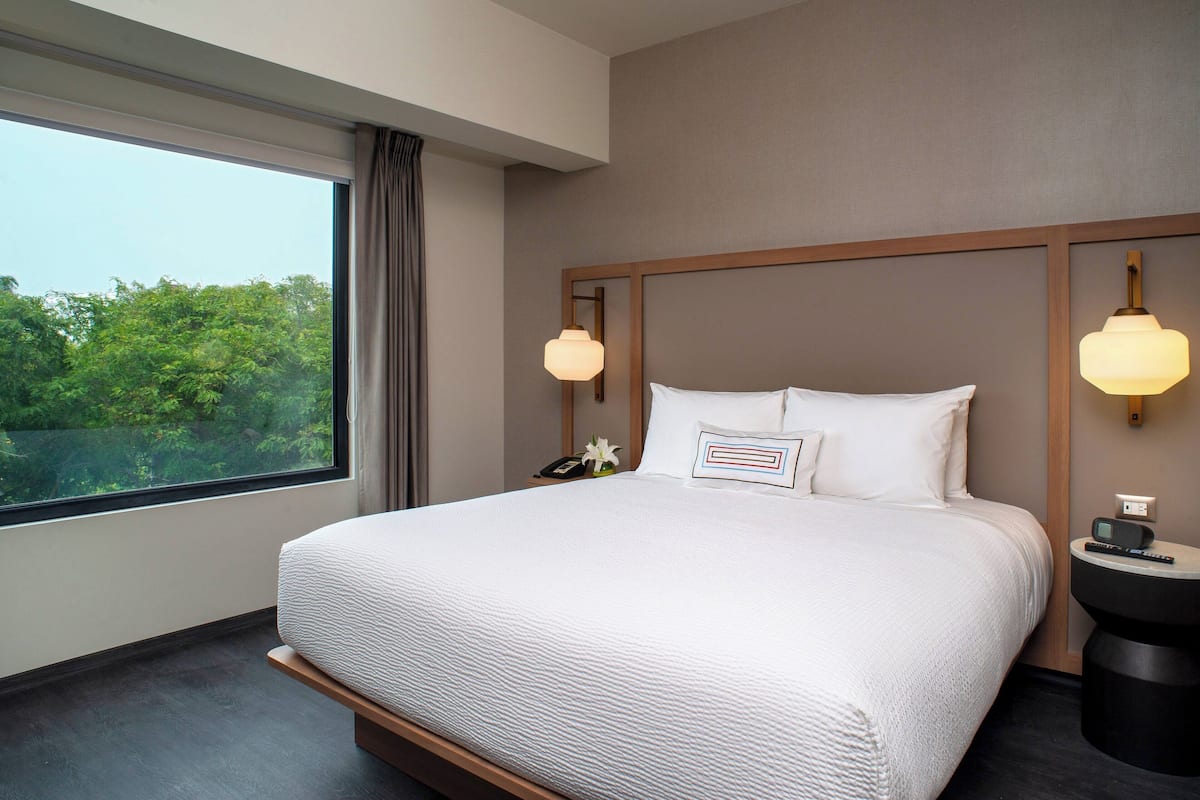 A hotel room with a large bed, a nightstand, a lamp, and a view of trees through the window.