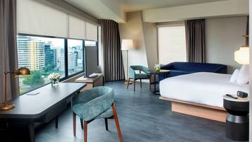 Habitación de hotel moderna con una cama grande, un escritorio, una silla, una mesita y vistas a la ciudad a través de amplias ventanas.