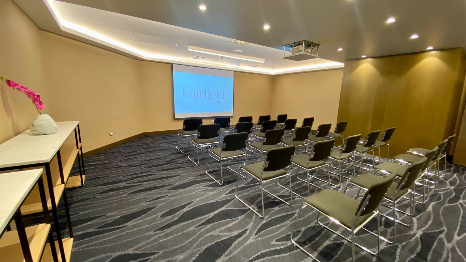 A conference room with a carpeted floor, rows of chairs, a projector screen, and a white table with a vase of flowers.