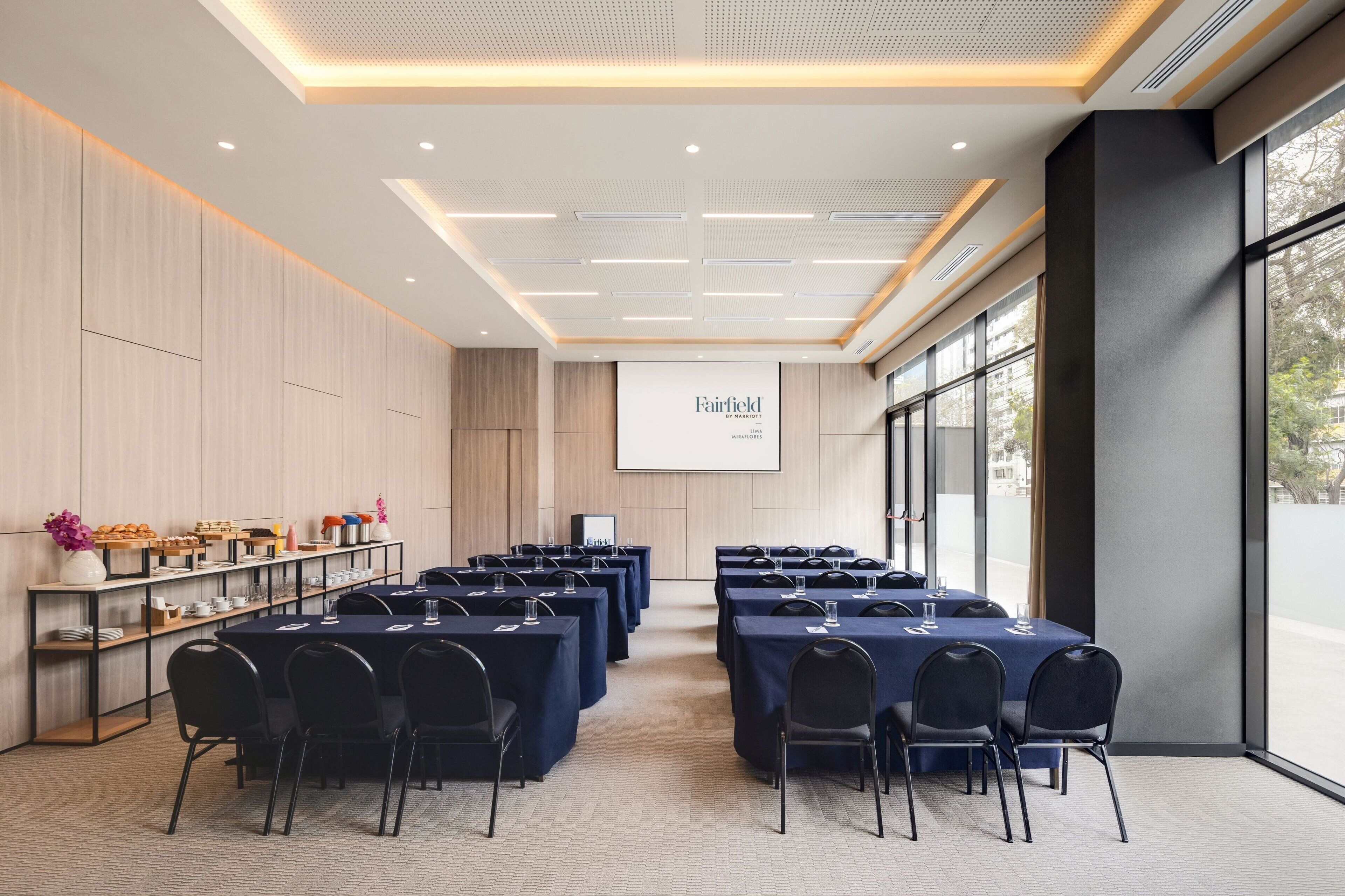 A conference room with tables and chairs, a projector screen, and a buffet area with food and drinks.