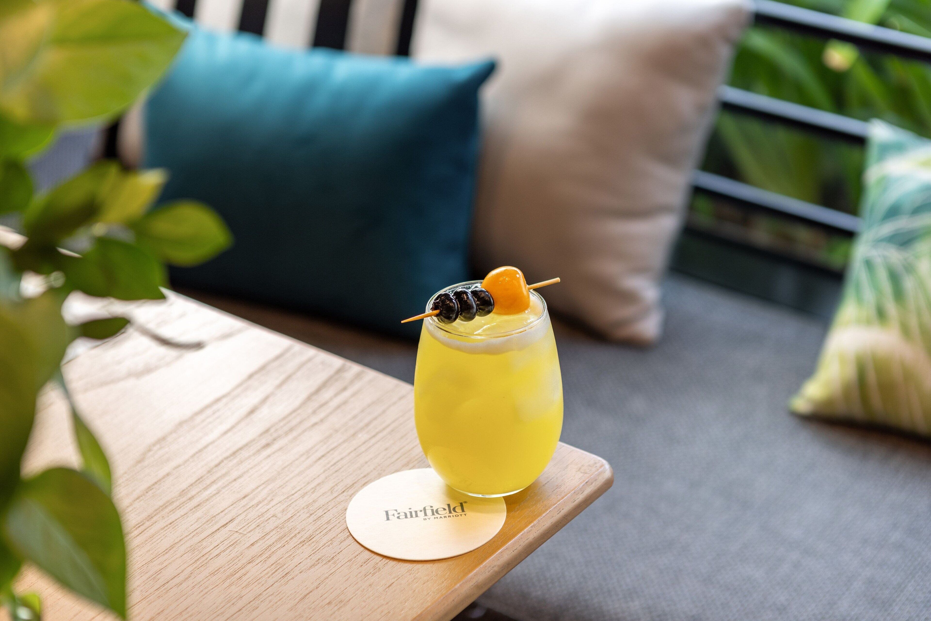 Un vaso de bebida amarilla sobre una mesa de madera con un posavasos con la etiqueta "Fairfield", en un entorno exterior tranquilo.
