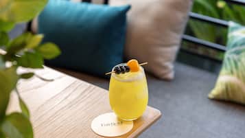 Un vaso de bebida amarilla sobre una mesa de madera con un posavasos con la etiqueta "Fairfield", en un entorno exterior tranquilo.