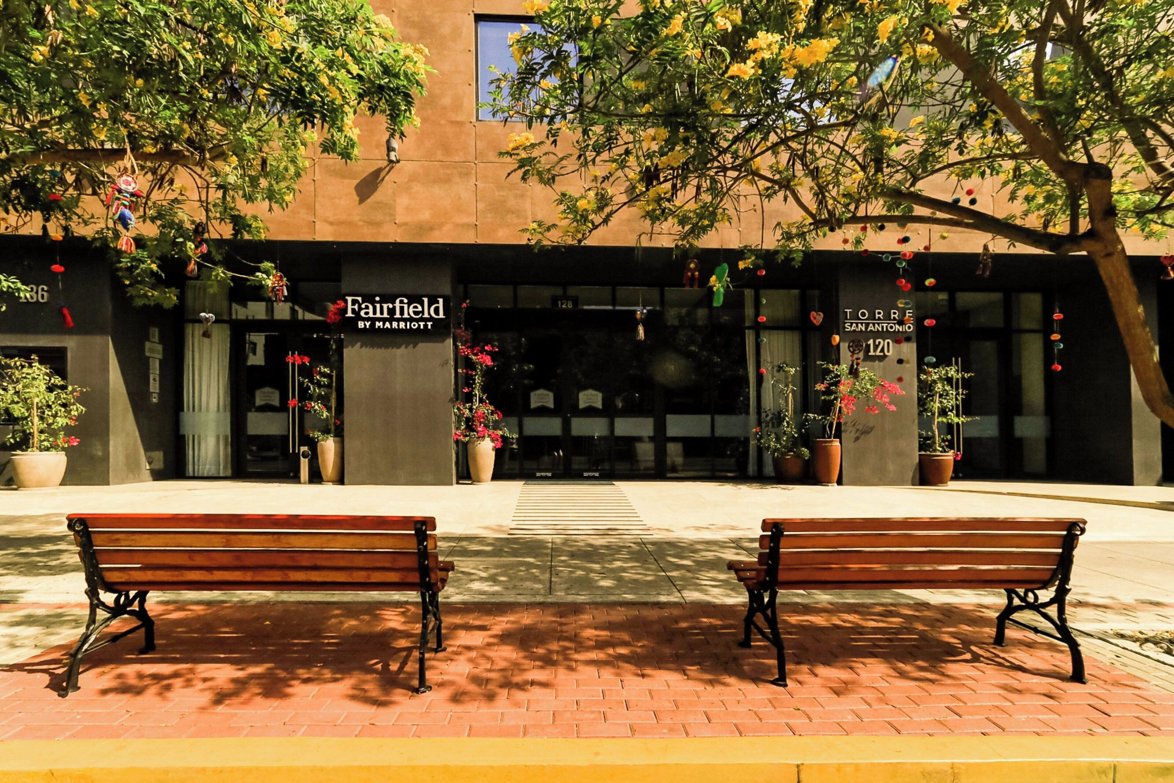 A hotel entrance with two benches in front, a sign for 'Fairfield by Marriott', and potted plants around the entrance.