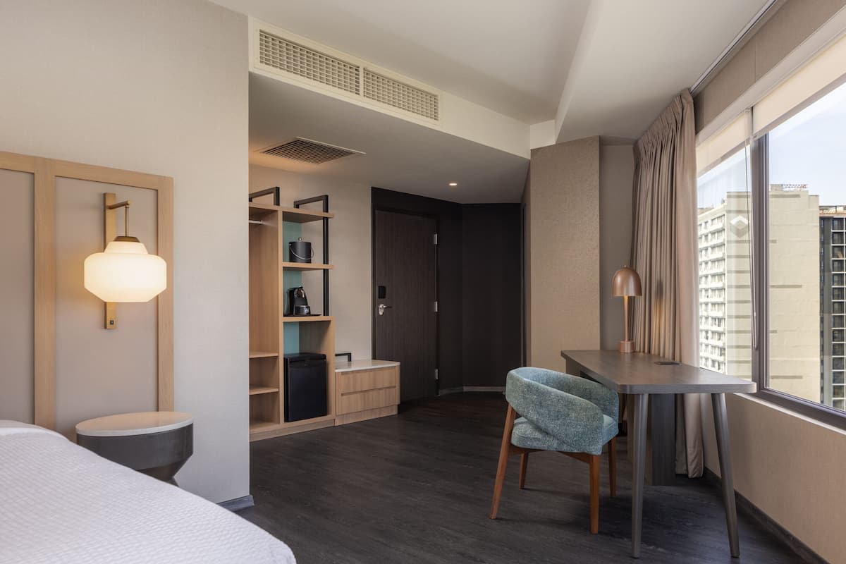 A modern hotel room with a bed, a desk, a chair, a wardrobe, and a large window offering a city view.