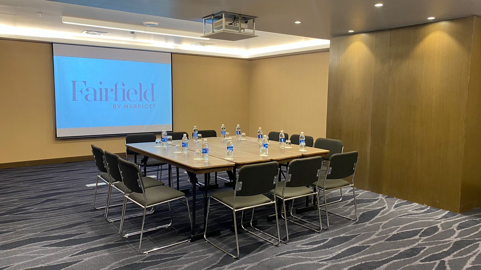 Un salón de conferencias con una pantalla grande que muestra 'Fairfield by Marriott', una mesa larga con sillas, botellas de agua y suelo alfombrado.
