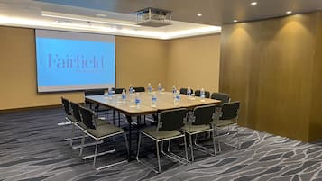 Un salón de conferencias con una pantalla grande que muestra 'Fairfield by Marriott', una mesa larga con sillas, botellas de agua y suelo alfombrado.
