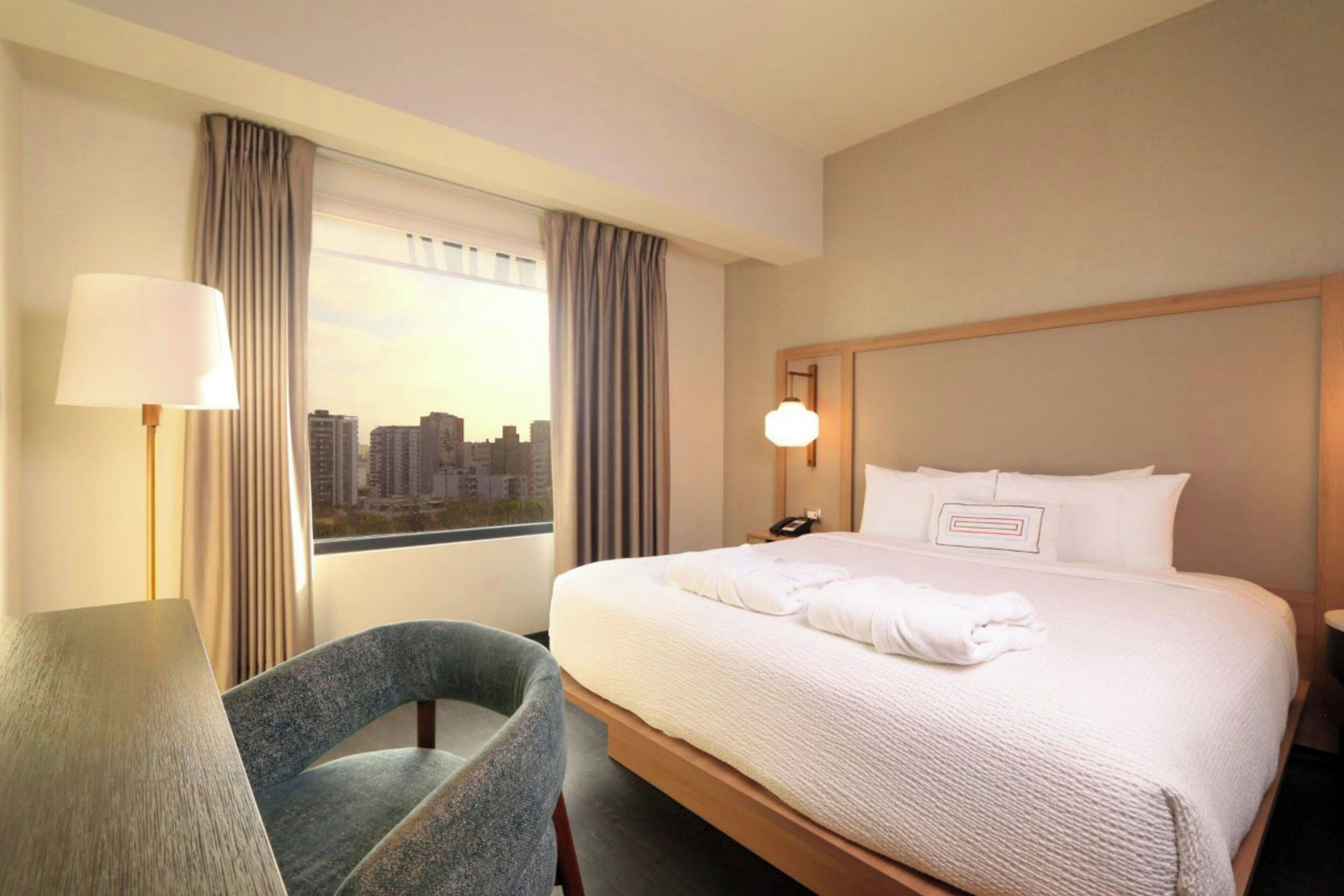 Habitación de hotel con una cama grande, una silla, un escritorio, una lámpara y vistas a la ciudad a través de la ventana.