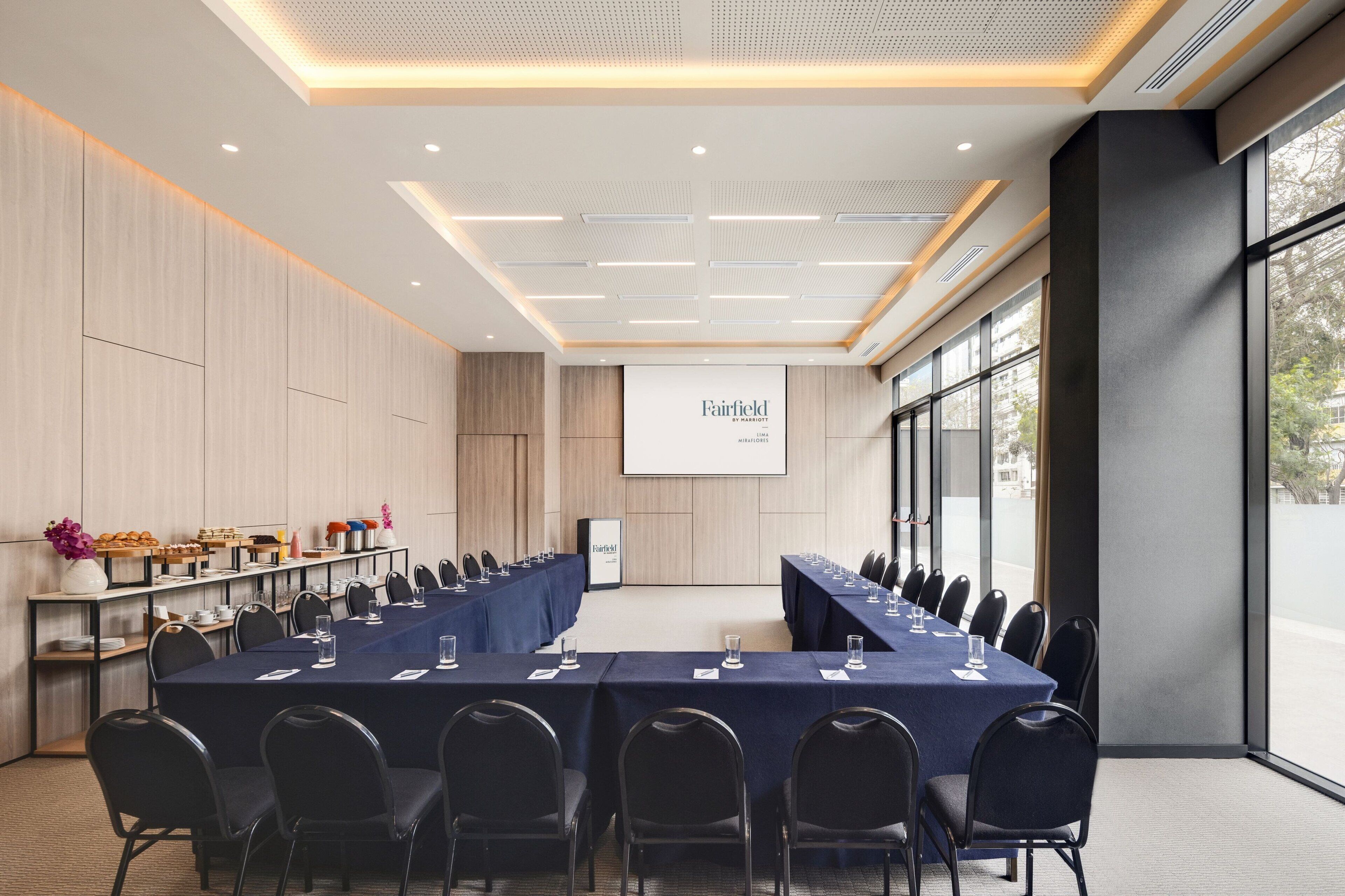 Un salón de conferencias con una larga mesa, sillas, una pantalla de proyector y una zona de buffet.