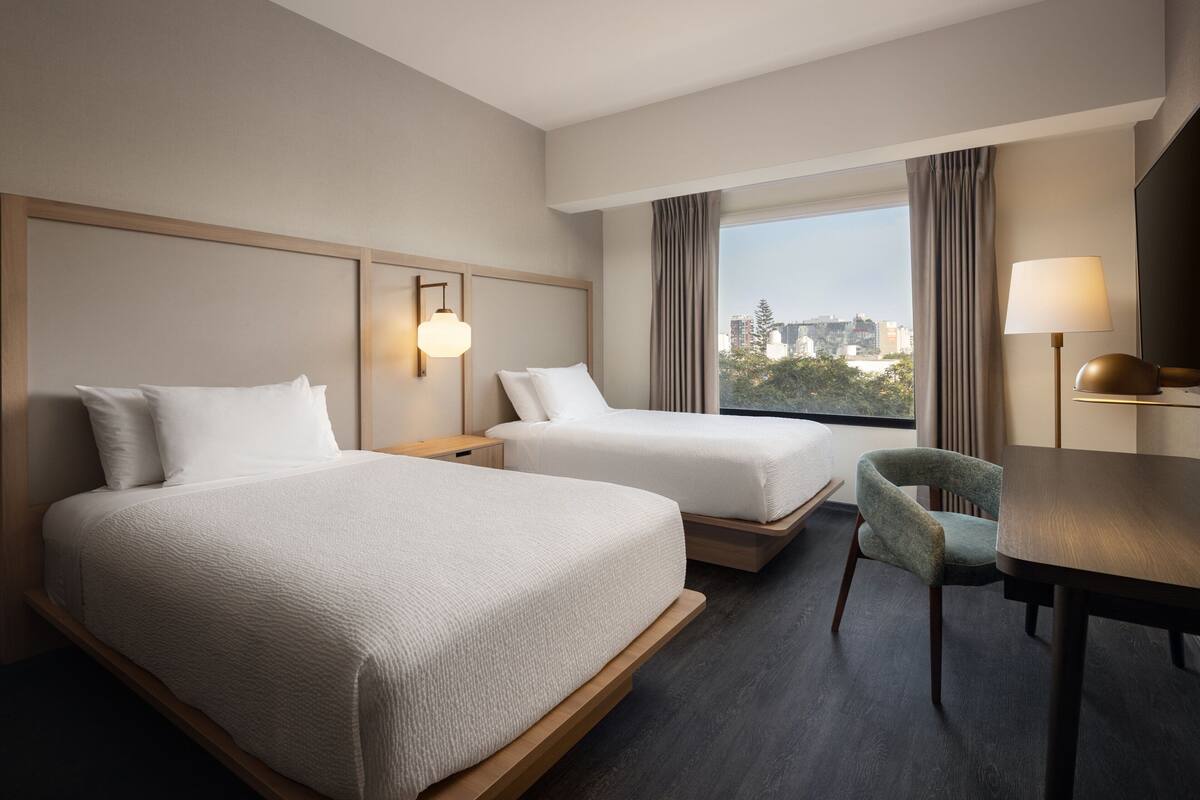 A hotel room with two beds, a desk, a chair, a window with curtains, and a lamp.