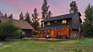 A log cabin with a deck and outdoor seating area, surrounded by a grassy yard and tall trees. - Cascade Getaway, Hot Tub, Fenced Acre, Pet Friendly (Bend)