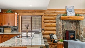 A log cabin interior with a kitchen island, stone fireplace, wooden flooring, and a view of the outdoors through a window. - Cascade Getaway, Hot Tub, Fenced Acre, Pet Friendly (Bend)