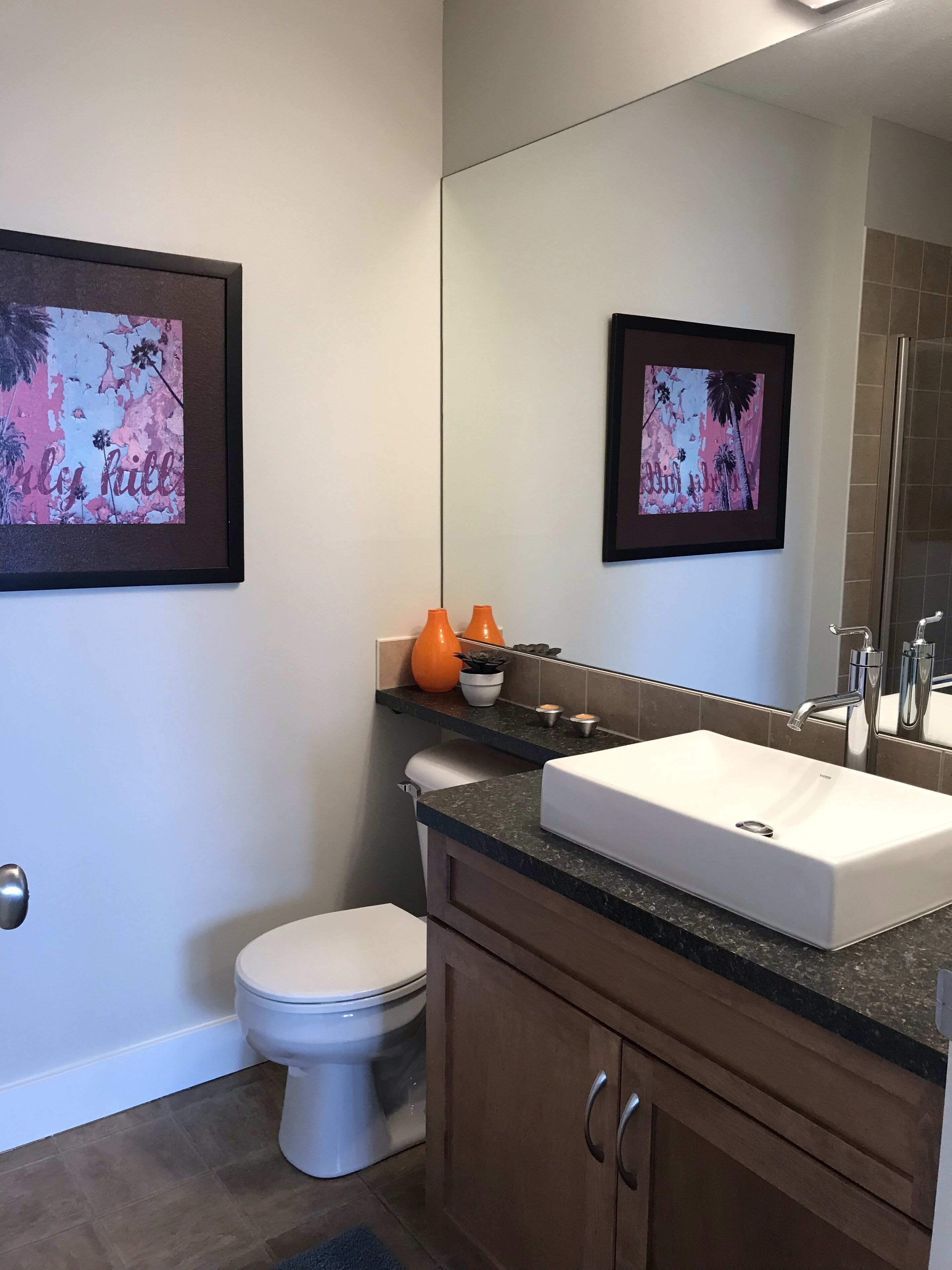 A bathroom with a white toilet, a white sink, and a mirror. There are two framed pictures on the wall, a small shelf with orange vases, and a potted plant.