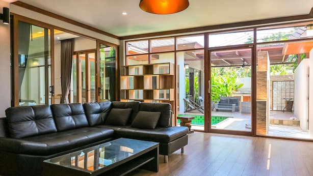 A modern living room with a black leather sofa, a glass coffee table, and a built-in bookshelf. - Nice House Pool Villa Phuket (Chalong)