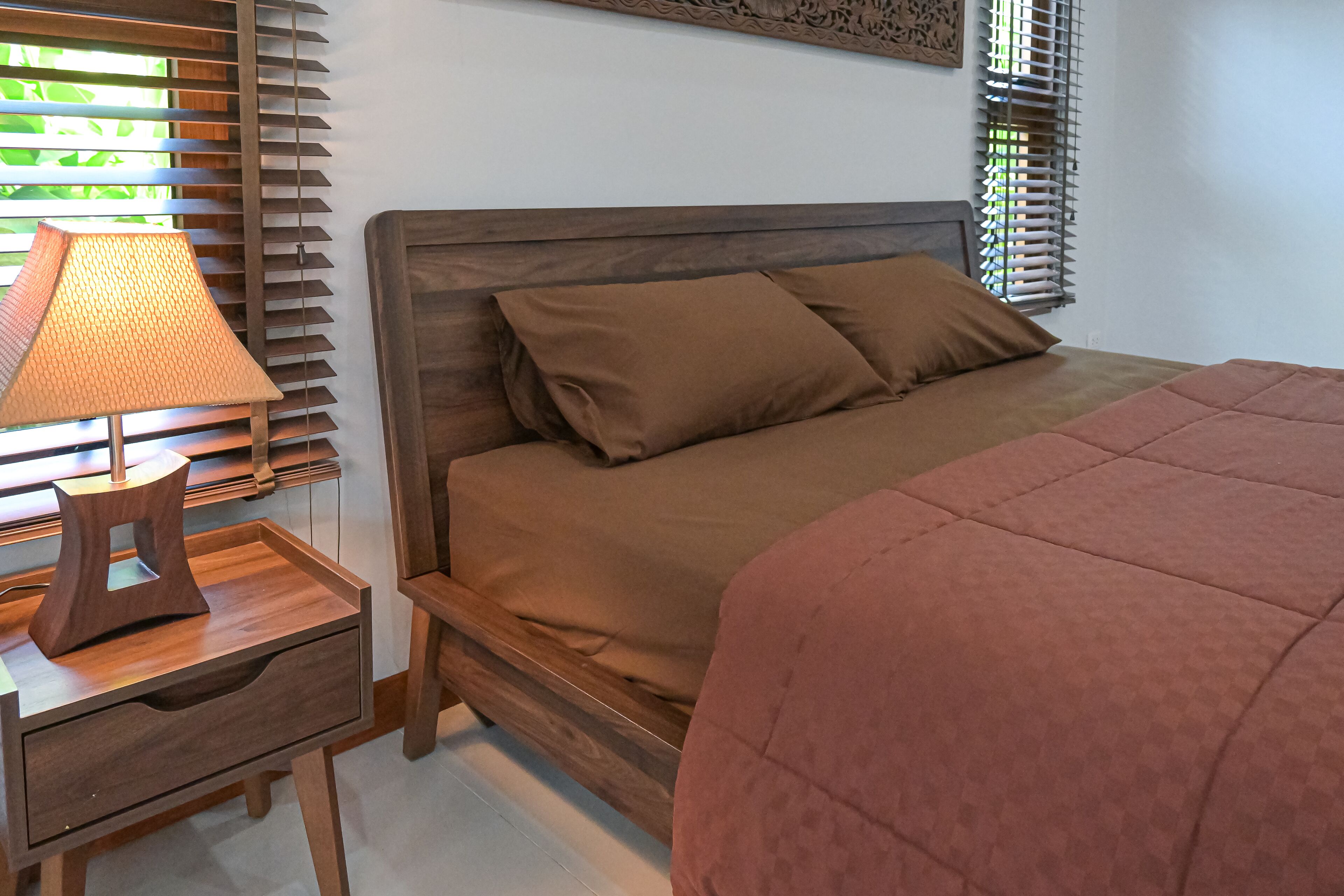 A bedroom with a wooden bed, brown bedding, a nightstand with a lamp, and wooden blinds.