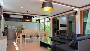 A modern living room with a black leather sofa, wooden dining table, and chairs. There is a kitchen area with a refrigerator and a fan. Large windows allow natural light to enter. - Nice House Pool Villa Phuket (Chalong)