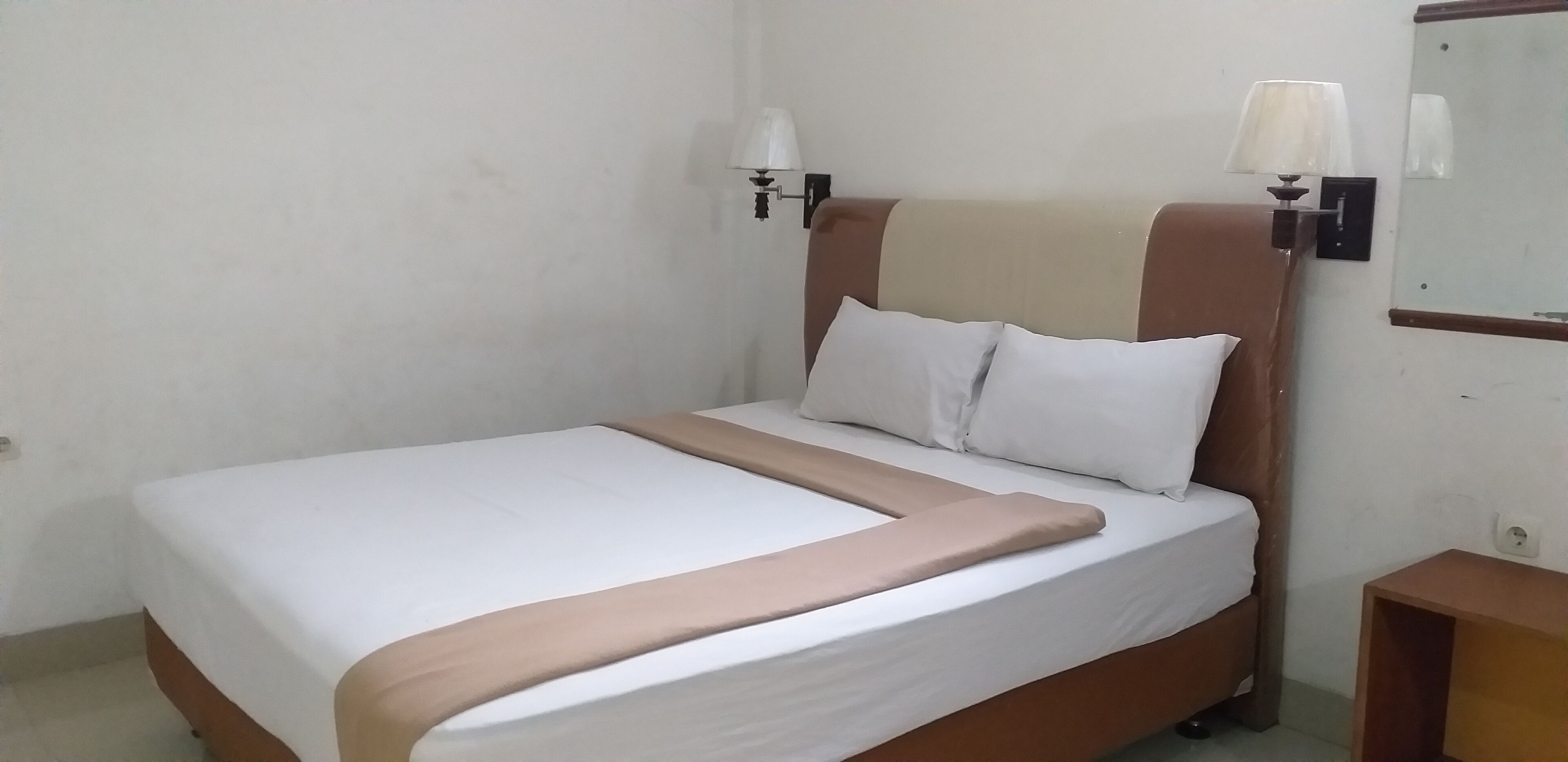 A neatly made bed with a white and beige bedspread, two pillows, and a wooden headboard. There is a nightstand with a lamp and a mirror on the wall beside the bed.