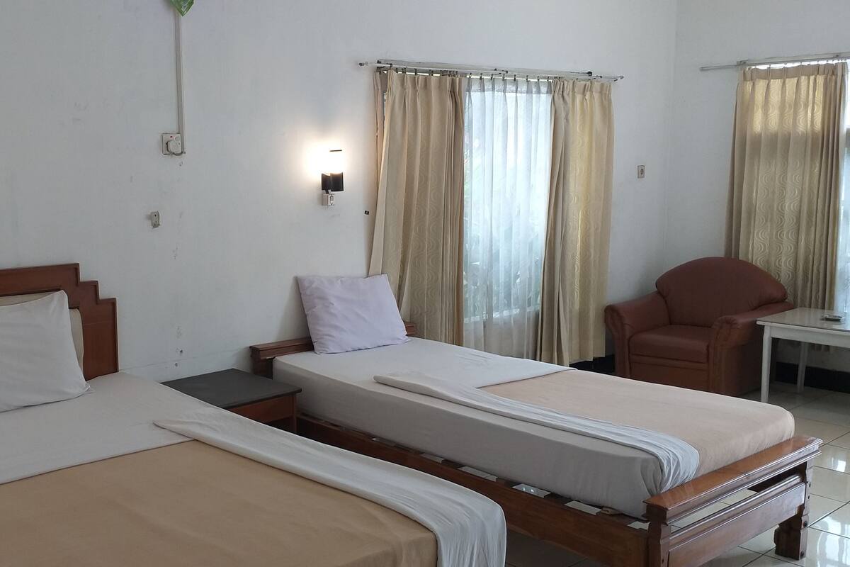Une chambre d’hôtel avec deux lits simples, une tête de lit en bois, une petite table, une chaise et une fenêtre avec des rideaux.