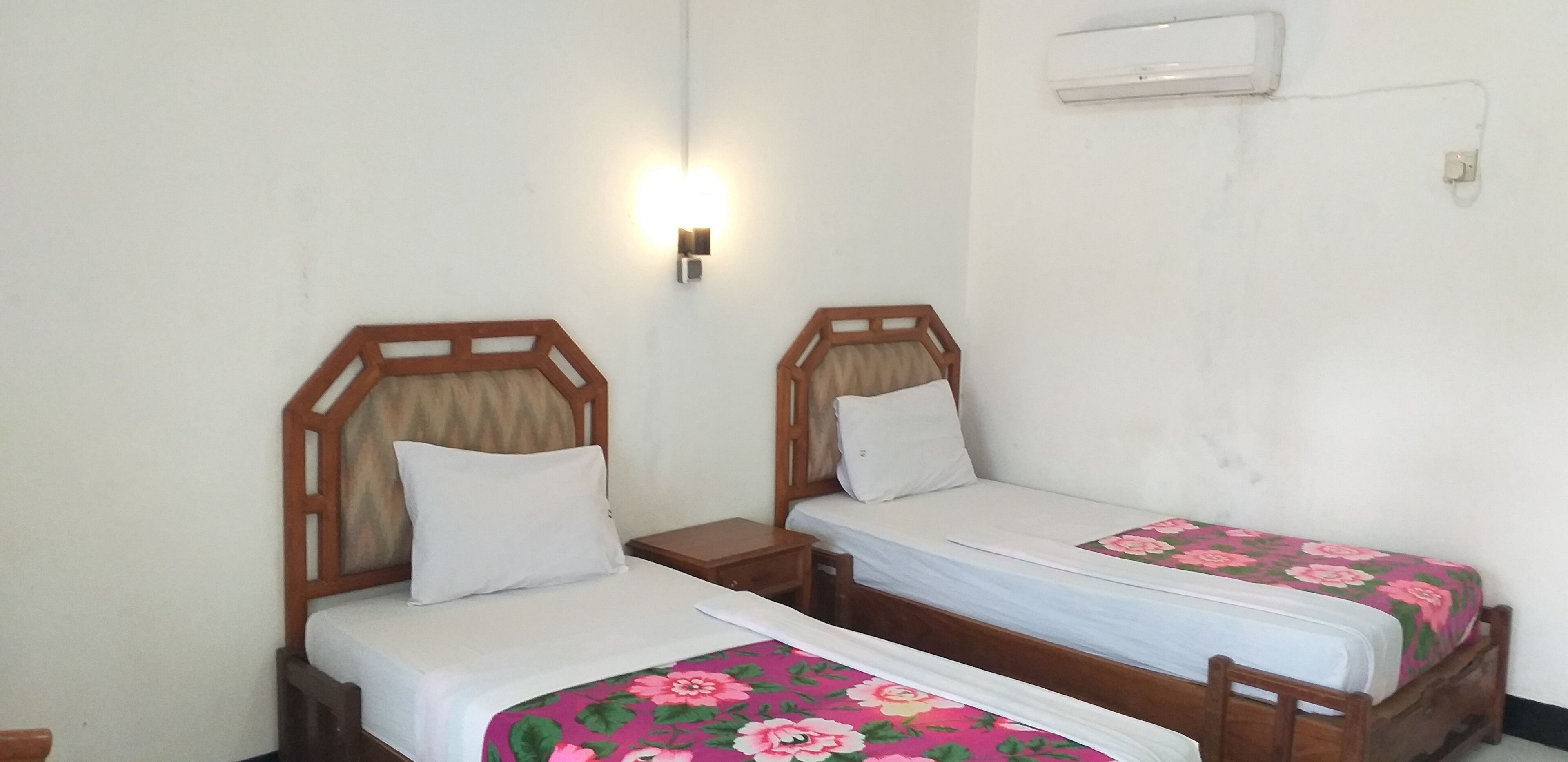 Two single beds with wooden headboards and white bedding, a wall-mounted air conditioner, and a small bedside table with a lamp.