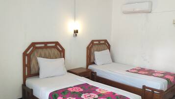 Two single beds with wooden headboards and white bedding, a wall-mounted air conditioner, and a small bedside table with a lamp.