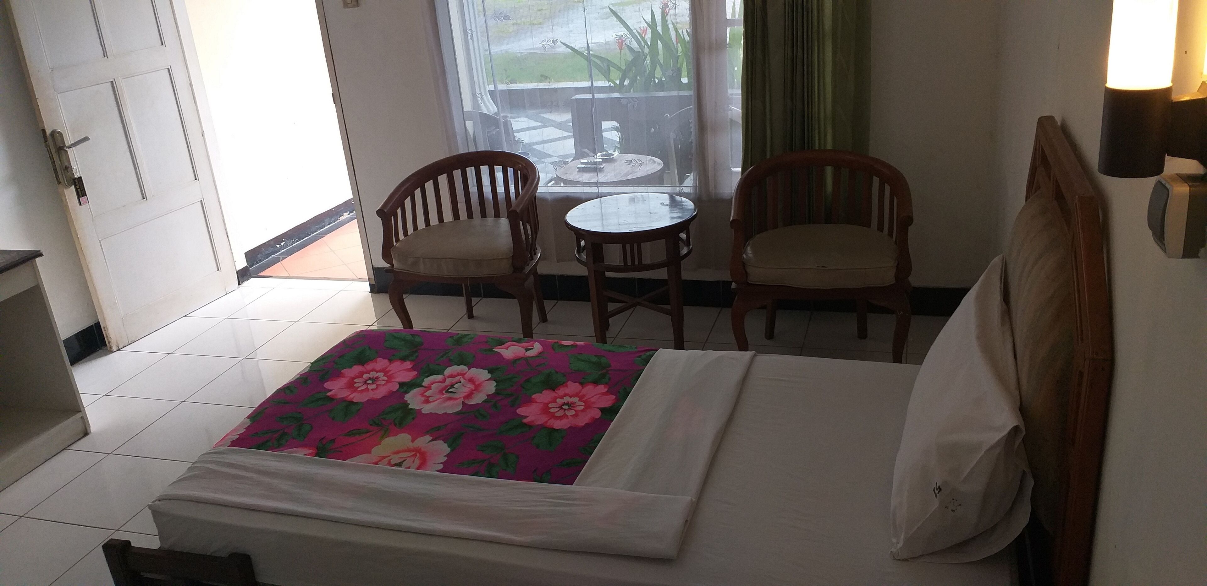A hotel room with a bed, two wooden chairs, a small table, and a view of the outdoors through a window.