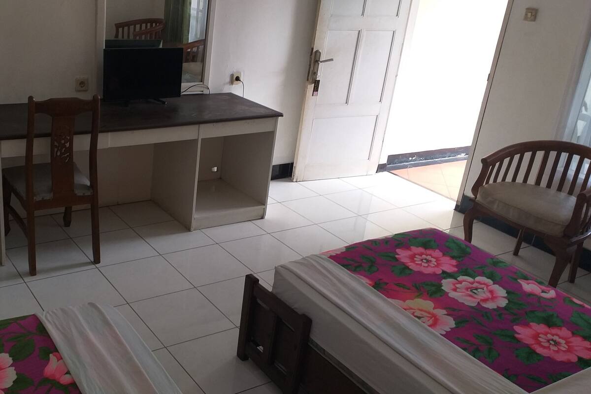 Une pièce comprenant un bureau, deux chaises, un lit recouvert d’une couvre-lit à motifs floraux et une porte.