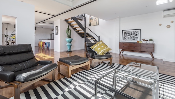 Un salon moderne avec un canapé en cuir noir, une table basse en bois et un tapis à rayures. Un escalier avec une rampe noire mène à un niveau supérieur.