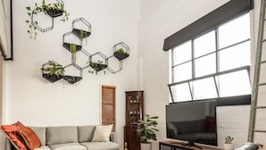 Un salon comprenant un canapé, un fauteuil en cuir noir, une table basse en bois, un téléviseur posé sur un meuble et des étagères murales hexagonales ornées de plantes.