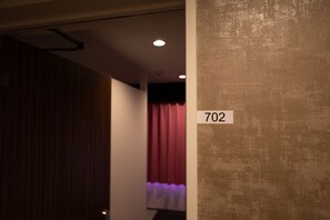 A hallway with a number sign '702' on a wall, a wooden door, and a curtain in the background. - Nikko Inn Minamisenba - Hostel (Osaka)