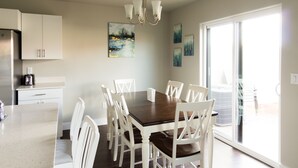 Un coin repas avec une table en bois et des chaises blanches, une cuisine avec des meubles blancs et des œuvres d’art accrochées aux murs.