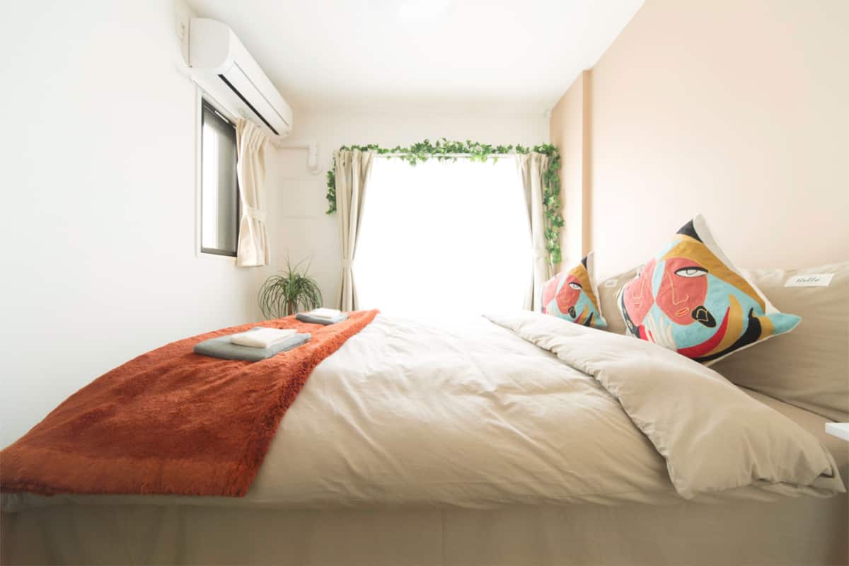 A bedroom with a bed, pillows, a blanket, an air conditioning unit, a window with curtains, and a plant.