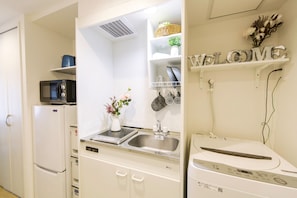 A kitchen with a refrigerator, microwave, sink, and a washing machine. There is a welcome sign on the wall and a basket on the shelf.