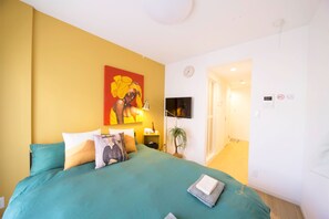 A bedroom with a teal bedspread, a painting on the wall, a clock, and a door leading to another room.
