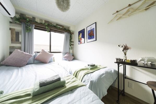 A bedroom with two beds, a bedside table with a 'WELCOME' sign, a window with curtains, and a wall-mounted air conditioner.