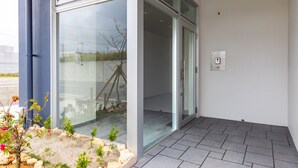 A modern building entrance with a glass door, a small garden area with plants, and a tiled floor. - Mr.KINJO in GRAND BLUE CHATAN (Chatan)