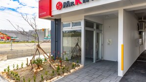 A Mr. Kinjo store with a modern entrance, a small garden area with plants and a wooden structure, and a clear sky. - Mr.KINJO in GRAND BLUE CHATAN (Chatan)