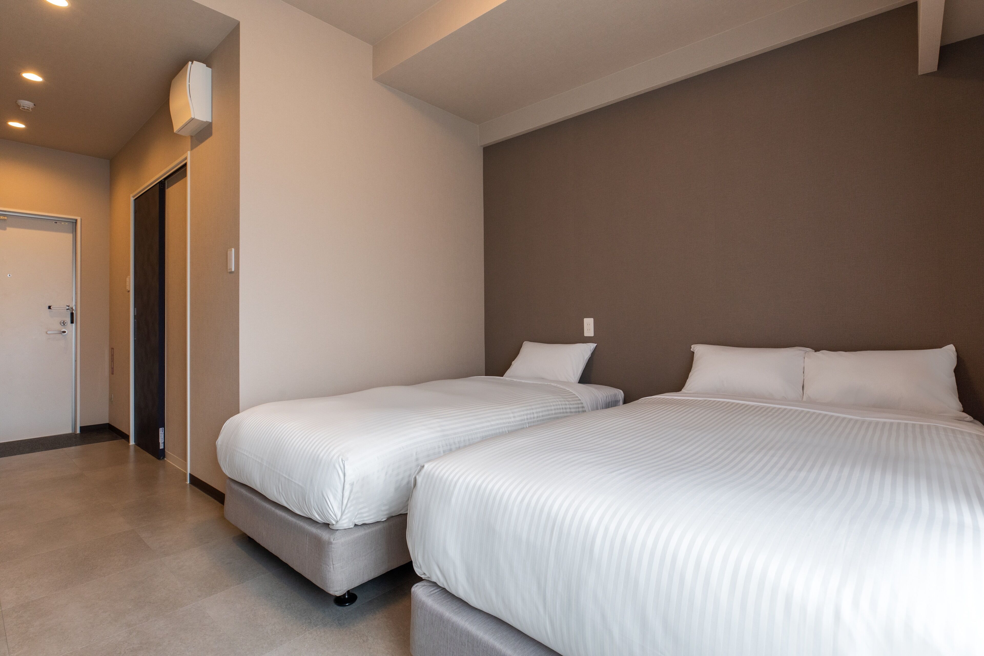 A hotel room with two single beds, a door, and a wall-mounted air conditioning unit.