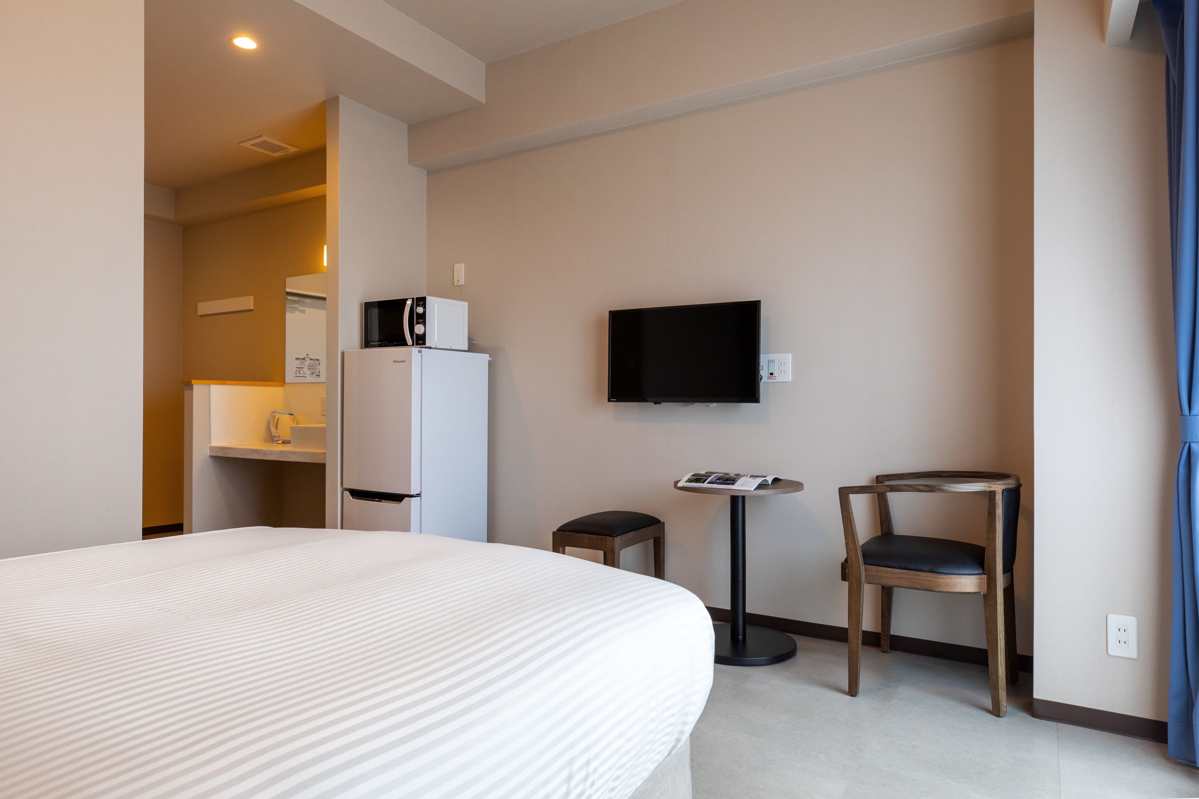 A hotel room with a large bed, a television mounted on the wall, a small table with a chair, and a refrigerator.