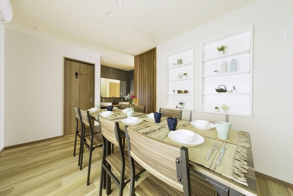 Un comedor con una larga mesa de madera dispuesta para cuatro personas, rodeada de sillas. Se aprecia una cocina al fondo con una puerta de madera y una encimera.