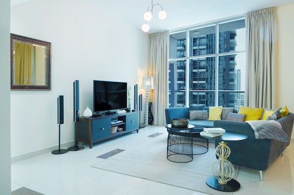 A modern living room with a large window, a flat-screen TV, a sofa with cushions, a coffee table, and a floor lamp.